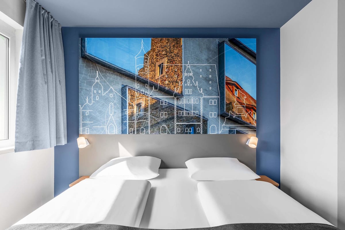 Une chambre d’hôtel avec un lit, une fenêtre avec des rideaux et un mur orné d’une œuvre d’art représentant un paysage urbain.