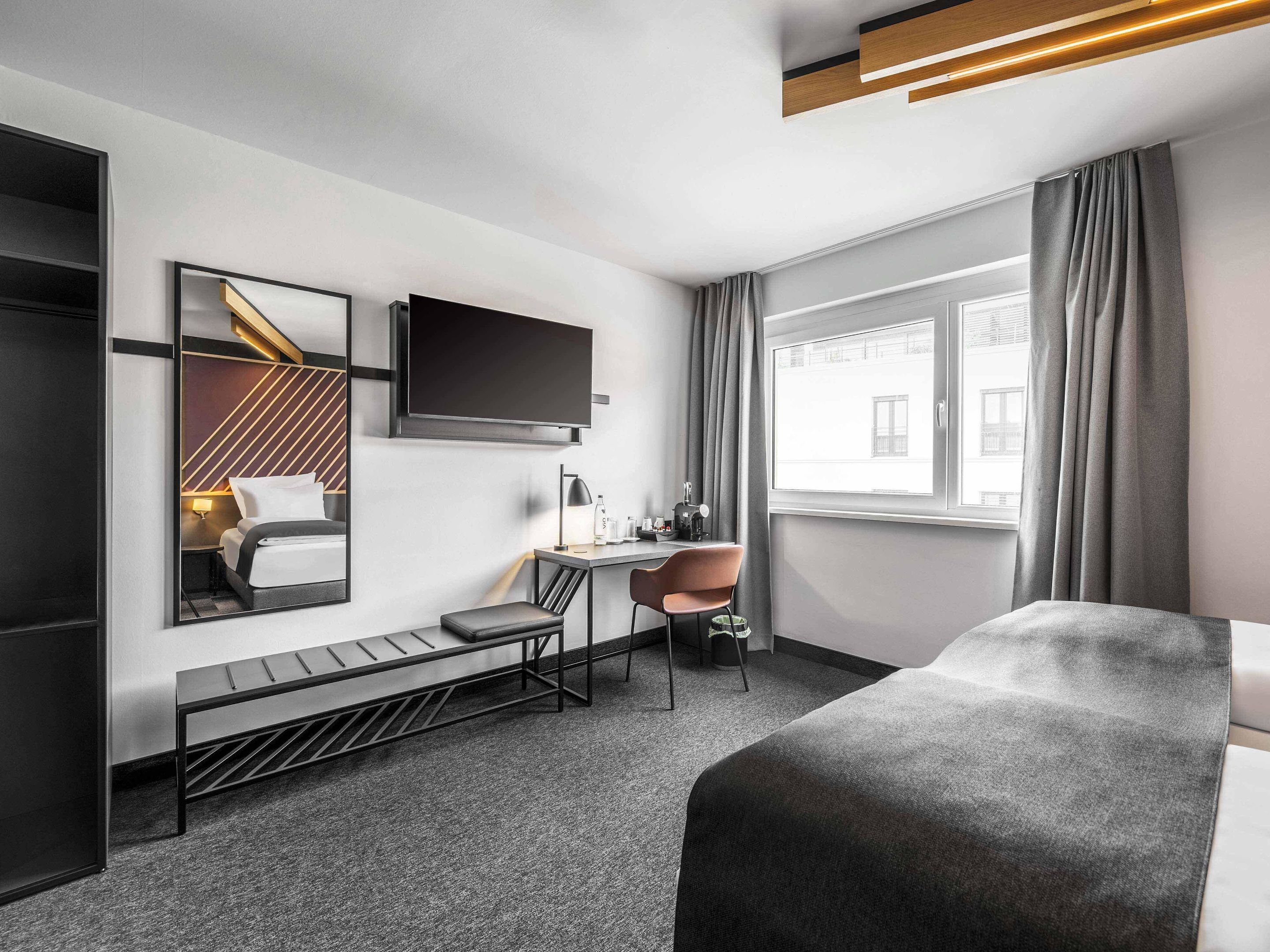 Ein modernes Hotelzimmer mit Bett, Schreibtisch mit Stuhl, Fernseher und großem Fenster mit Vorhängen.