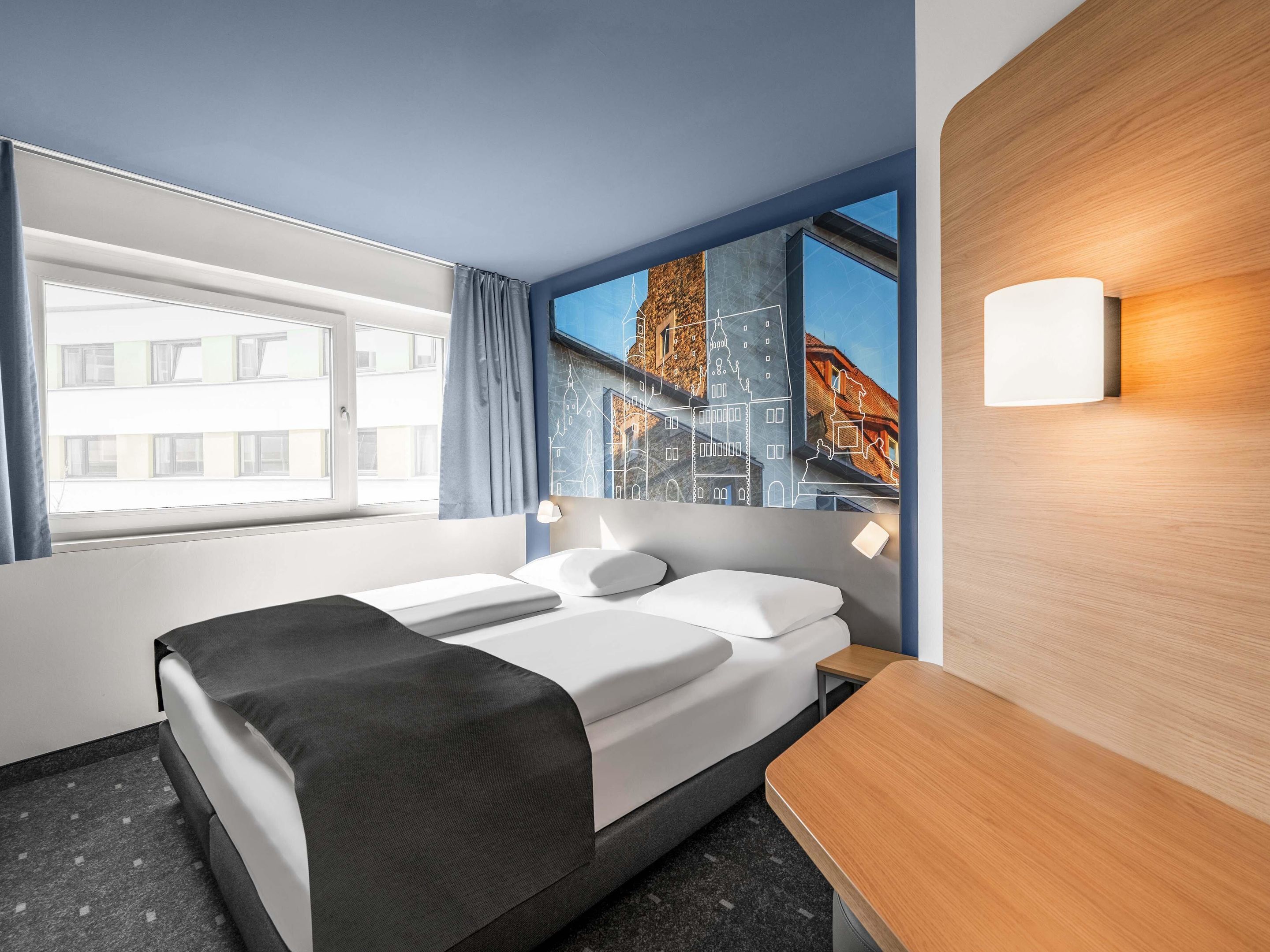 Ein Hotelzimmer mit Bett, Nachttisch, einem Fenster mit Vorhängen und einer Wandleuchte.
