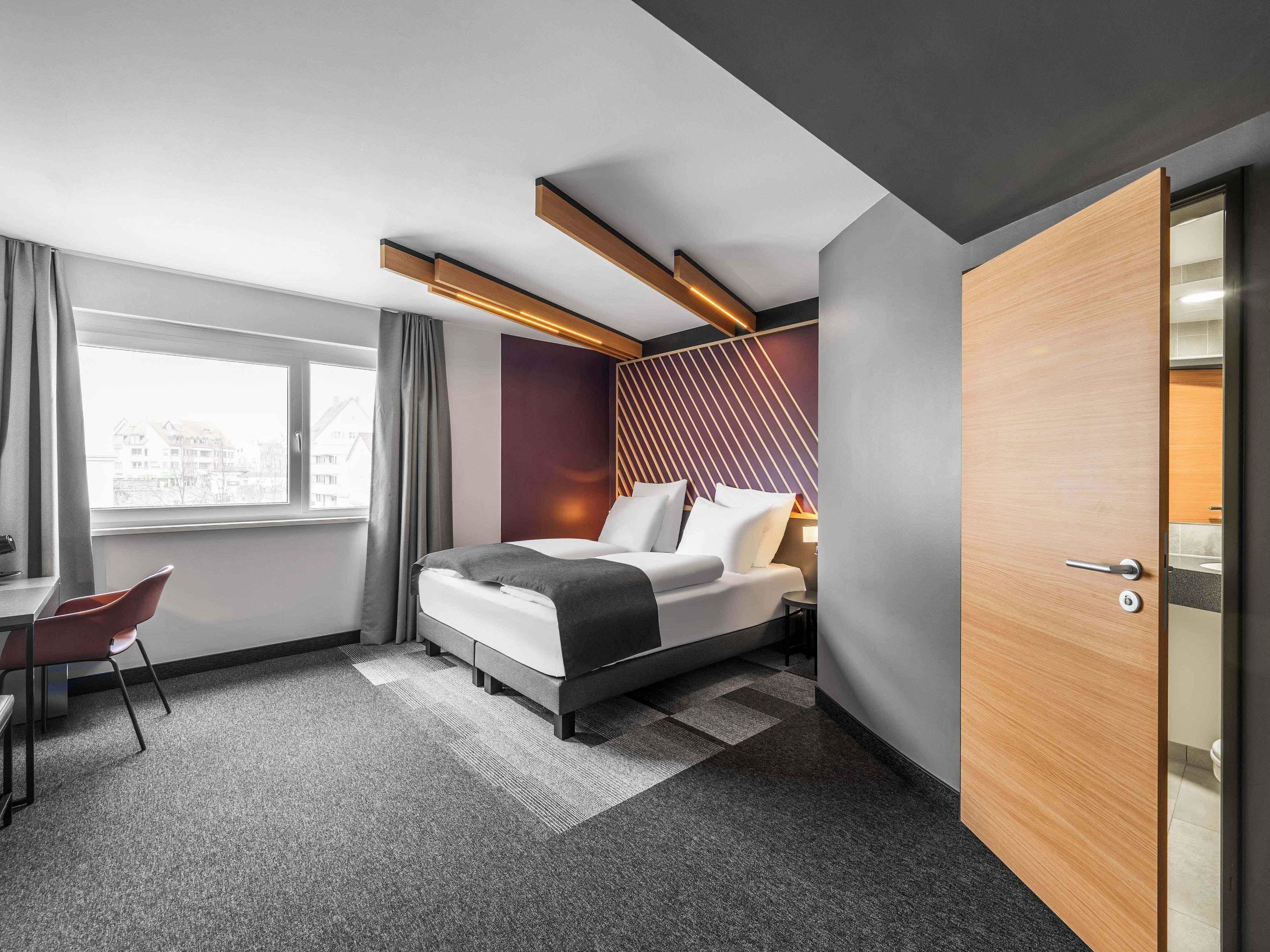 Ein modernes Hotelzimmer mit Bett, Schreibtisch und Blick auf Gebäude vor dem Fenster.