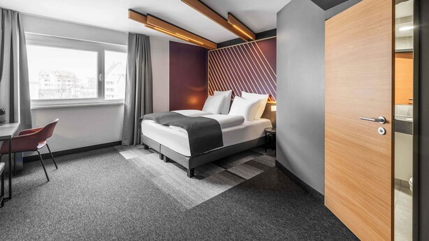 Une chambre d’hôtel moderne, équipée d’un lit, d’un bureau et offrant une vue sur les bâtiments à travers la fenêtre.