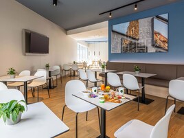 Un comedor moderno con sillas y mesas blancas, un cuadro grande de un paisaje urbano y un televisor montado en la pared.