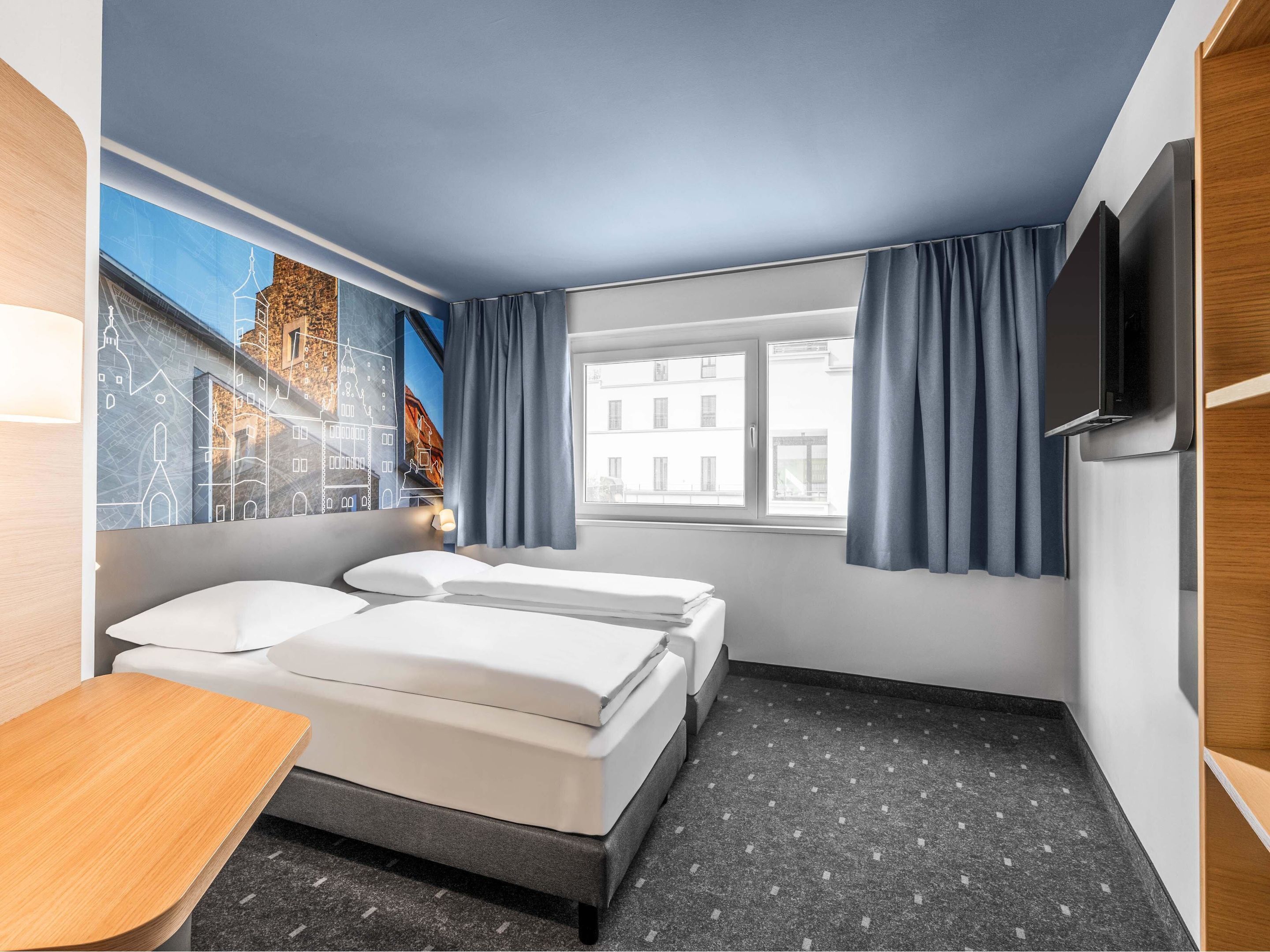 A hotel room with two beds, a desk, a TV, and a window with curtains.