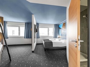 Ett hotellrum med en säng, ett skrivbord, en stol, ett fönster med gardiner och en duschhörna.