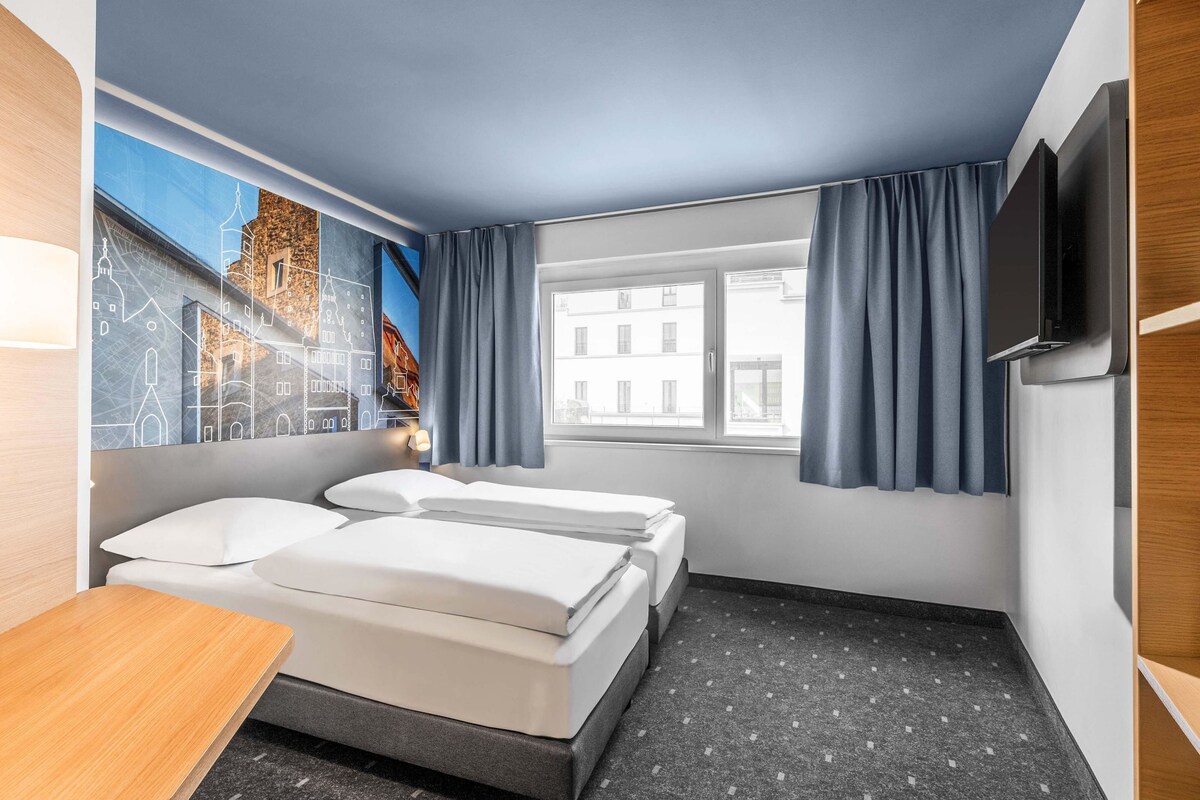 A hotel room with two beds, a desk, a TV, and a window with curtains.