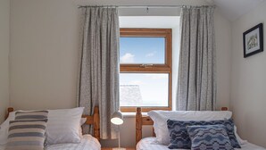 Une chambre avec deux lits simples, une table de chevet avec une lampe, une fenêtre avec des rideaux et un tableau encadré au mur.