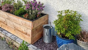 Un bac à fleurs en bois contenant diverses plantes, un arrosoir en métal et une pancarte indiquant « Corner House ».