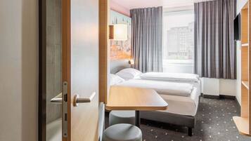 Quarto de hotel com cama, escrivaninha e cadeira. Há uma janela com cortinas, uma luminária e vista para edifícios.
