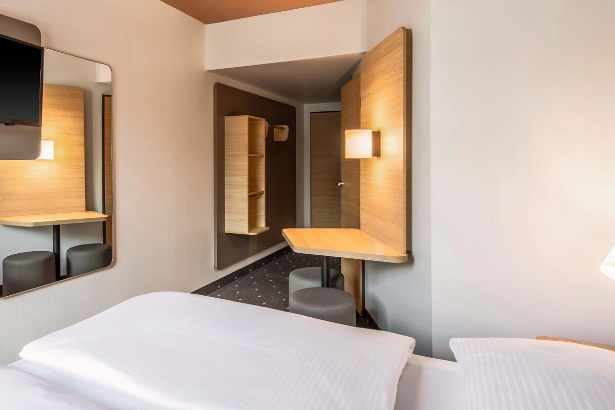 A hotel room with a large bed, a desk, a chair, a mirror, and a wall-mounted shelf.
