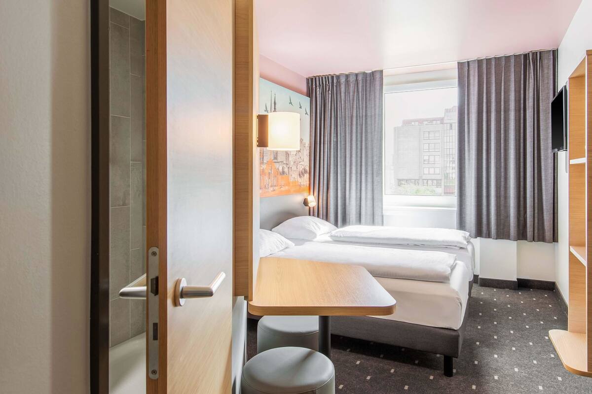 A hotel room with a bed, a desk, and a chair. There is a window with curtains, a lamp, and a view of buildings outside.