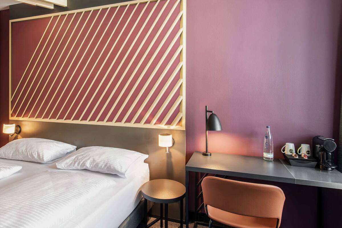A hotel room with a bed, bedside lamps, a desk with a chair, and a water bottle.