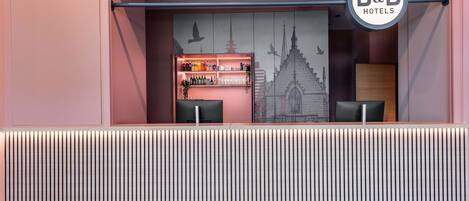 Un espace d’accueil avec une enseigne B&B Hotels, un bar avec diverses bouteilles et une fresque représentant une église.