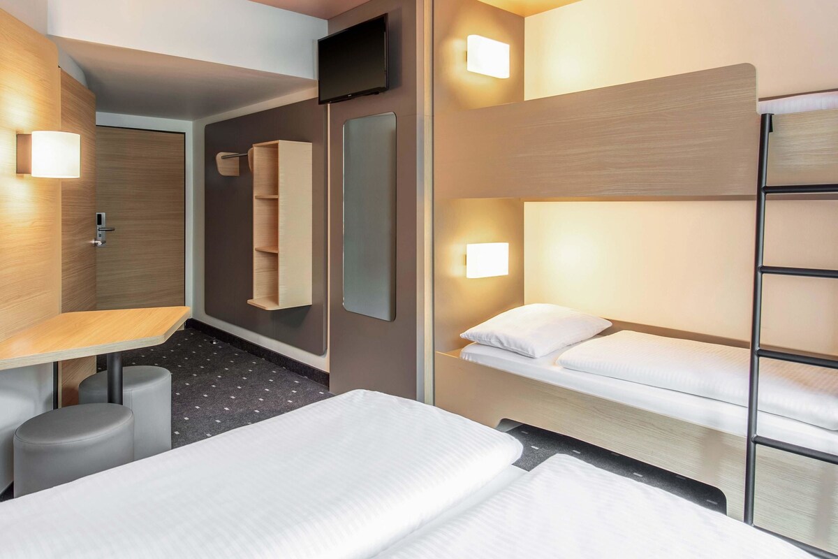 A hotel room with a bunk bed, a desk with a chair, a mirror, and a wall-mounted TV.