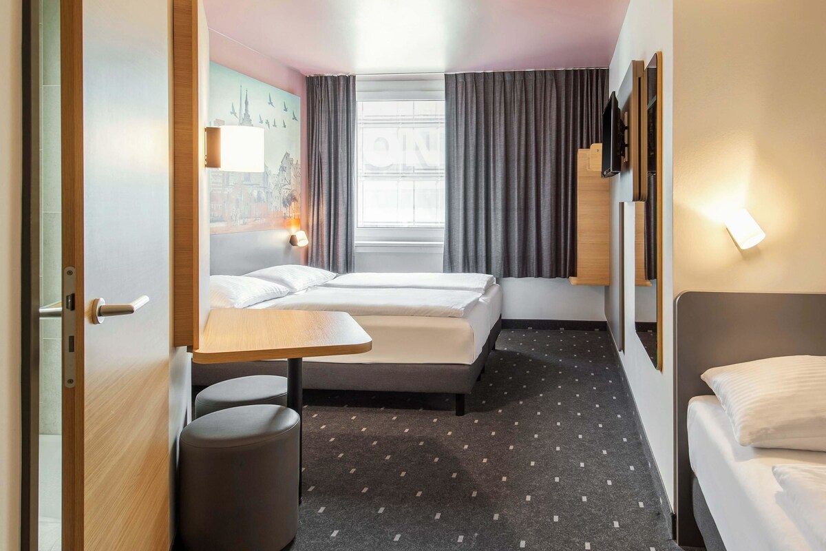 A hotel room with two beds, a small table, a chair, a wall-mounted TV, and a window with curtains.