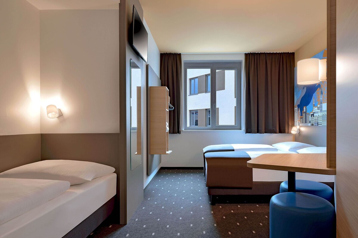 Ein Hotelzimmer mit Bett, Schreibtisch, Stuhl, einem Fenster mit Vorhängen und einem an der Wand befestigten Fernseher.
