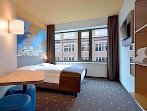 Habitación de hotel con cama, escritorio, silla, televisor y ventana con vista a la ciudad.