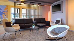 Een moderne woonkamer met een modulaire bank, een witte fauteuil, een ronde stoel en een salontafel. Er is een groot raam met zonwering, een flatscreen televisie aan de muur bevestigd en een decoratief wandobject.