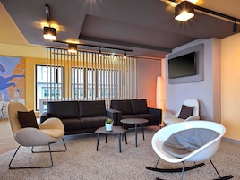 Una sala de estar moderna con un sofá modular, un sillón blanco, un asiento redondo y una mesa de centro. Hay un ventanal con persianas, un televisor de pantalla plana montado en la pared y un adorno en la pared.