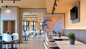 A modern dining area with multiple tables and chairs, a large mural on the wall, and a TV mounted above a seating area. - B&B Hotel Hannover-City (Hannover)