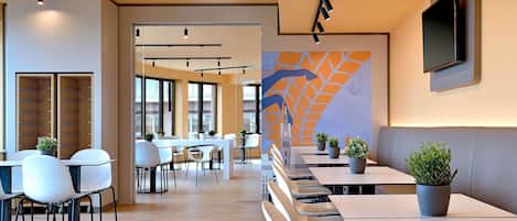 A modern dining area with multiple tables and chairs, a large mural on the wall, and a TV mounted above a seating area.
