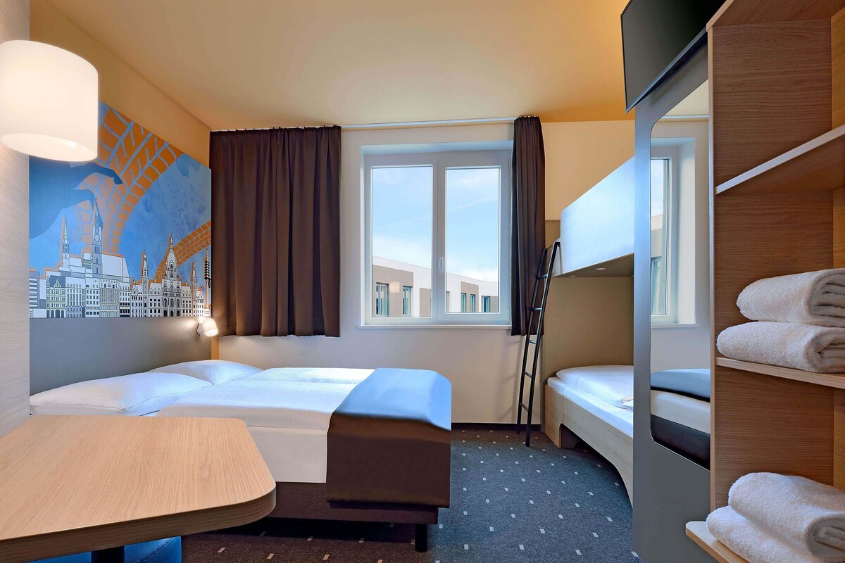 Ein Hotelzimmer mit zwei Betten, einem Schreibtisch, einem Stuhl, einem Fenster mit Vorhängen, einem Wandbild und einer Ablage mit Handtüchern.
