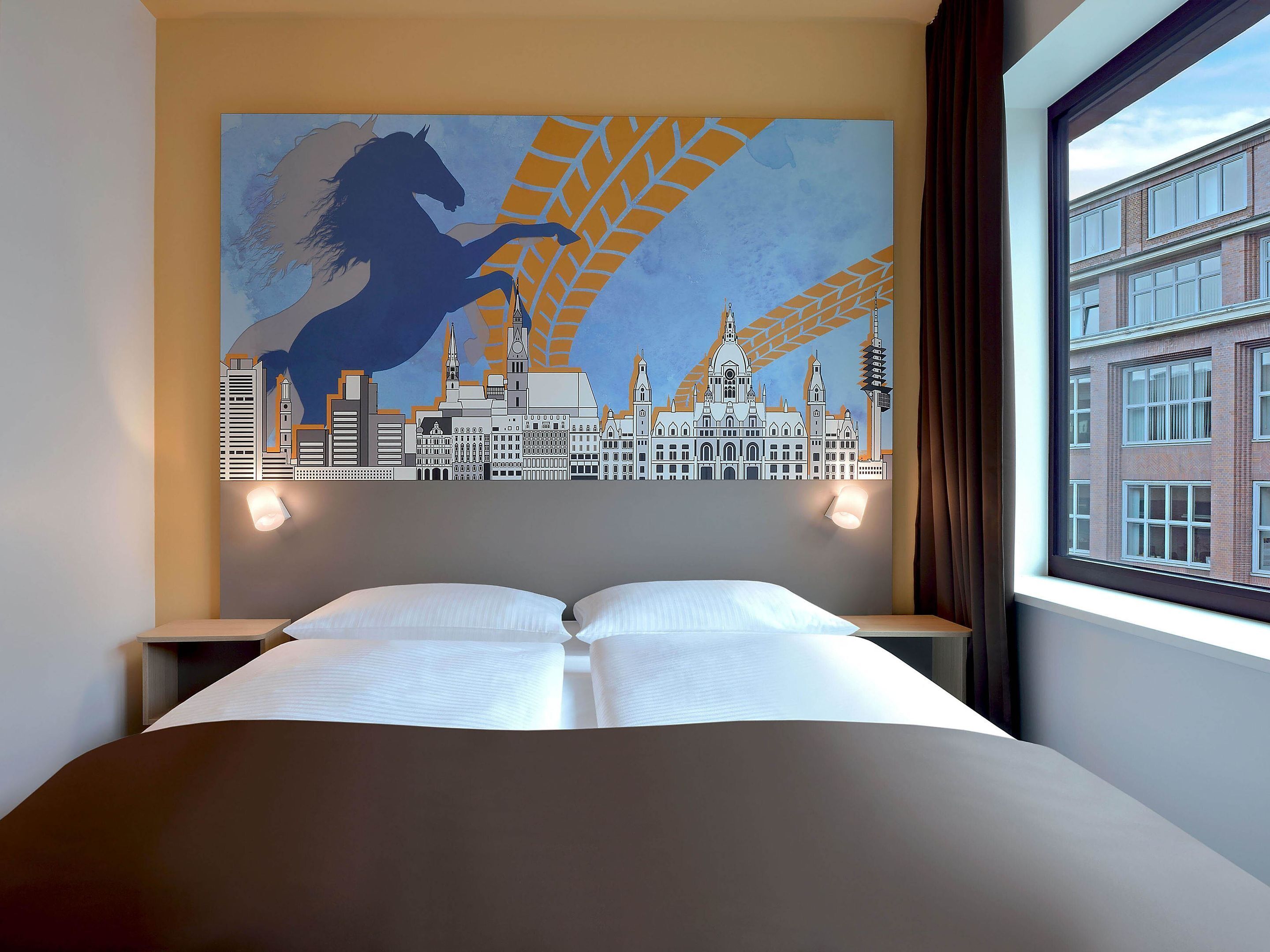A hotel room with a bed, two bedside tables, a cityscape mural on the wall, and a window with a view of buildings.