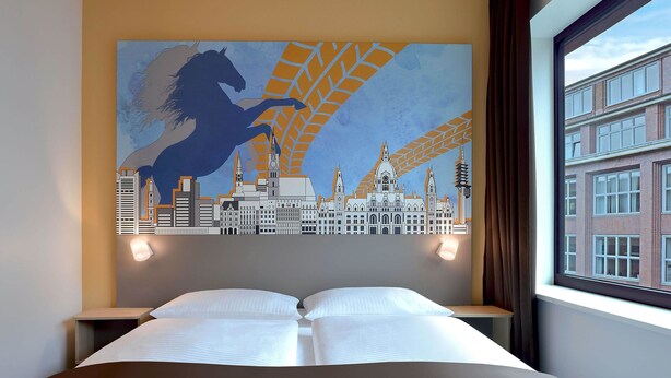 A hotel room with a bed, two bedside tables, a cityscape mural on the wall, and a window with a view of buildings. - B&B Hotel Hannover-City (Hannover)