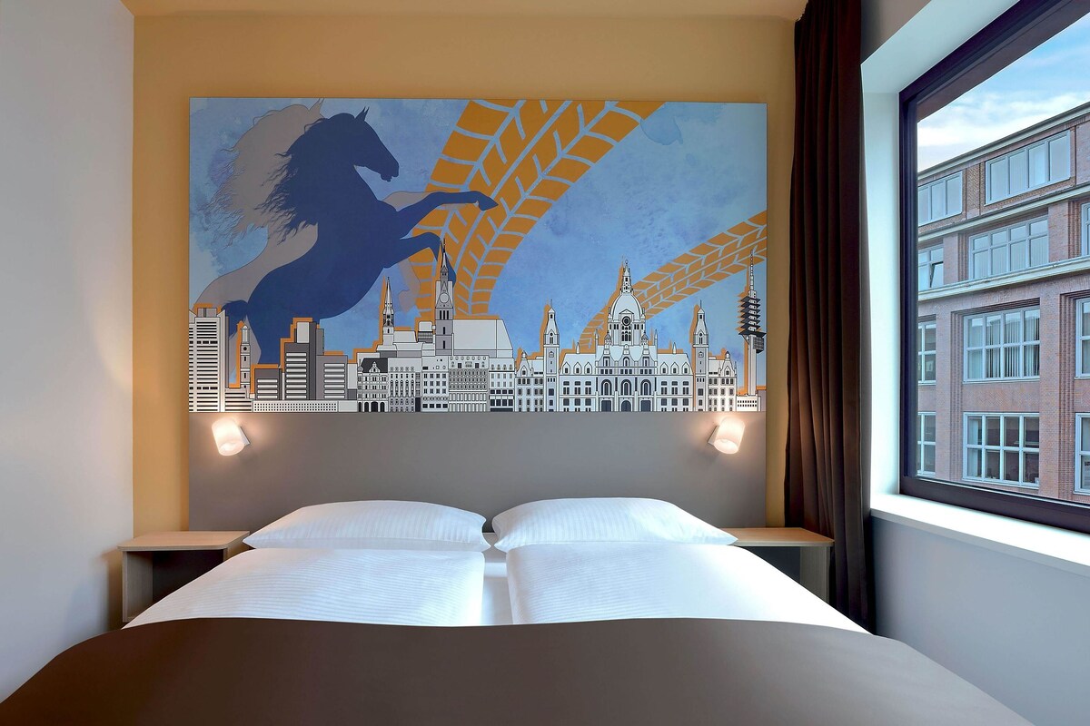 Ein Hotelzimmer mit einem Bett, zwei Nachttischen, einem Stadtbild-Wandgemälde und einem Fenster mit Blick auf Gebäude.