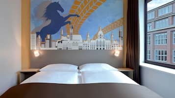 A hotel room with a bed, two bedside tables, a cityscape mural on the wall, and a window with a view of buildings.