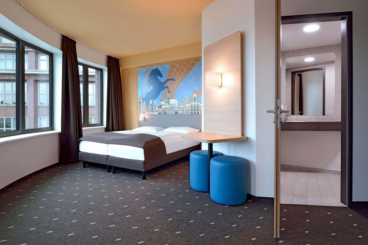 Ein Hotelzimmer mit Bett, Schreibtisch, zwei blauen Hocker, einem Stadtbild-Wandgemälde und einem durchgehenden Türrahmen sichtbaren Badezimmer.