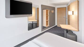 A modern hotel room with a bed, a TV mounted on the wall, a wooden wardrobe, and a small desk with a chair.