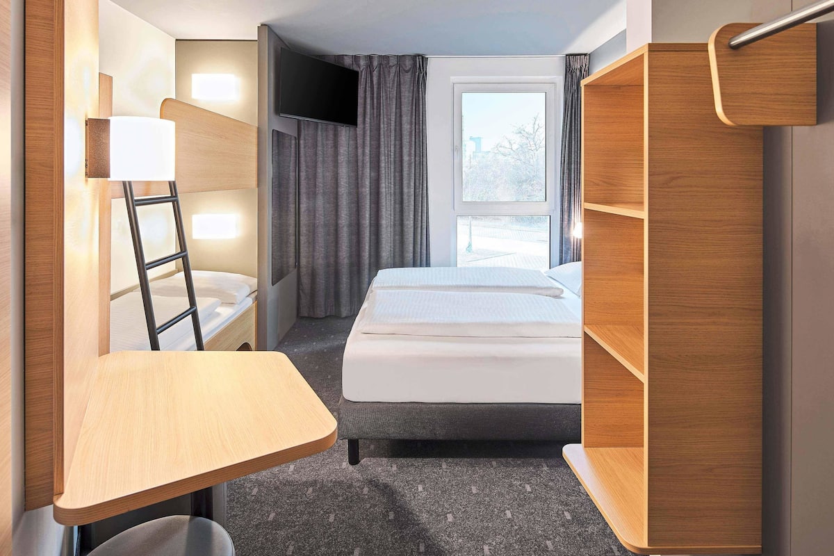 A compact hotel room with a bed, a desk, a chair, a TV, and a window with curtains.
