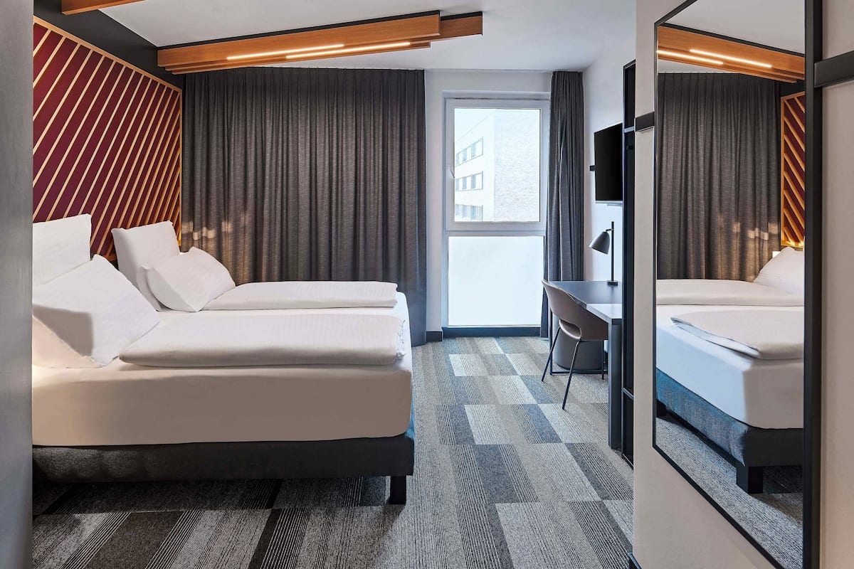 A modern hotel room with two beds, a desk, and a large window.