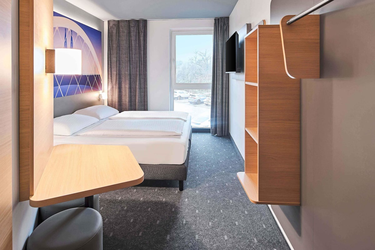 A modern hotel room with a bed, a desk, a chair, a TV, and a view of the outside.