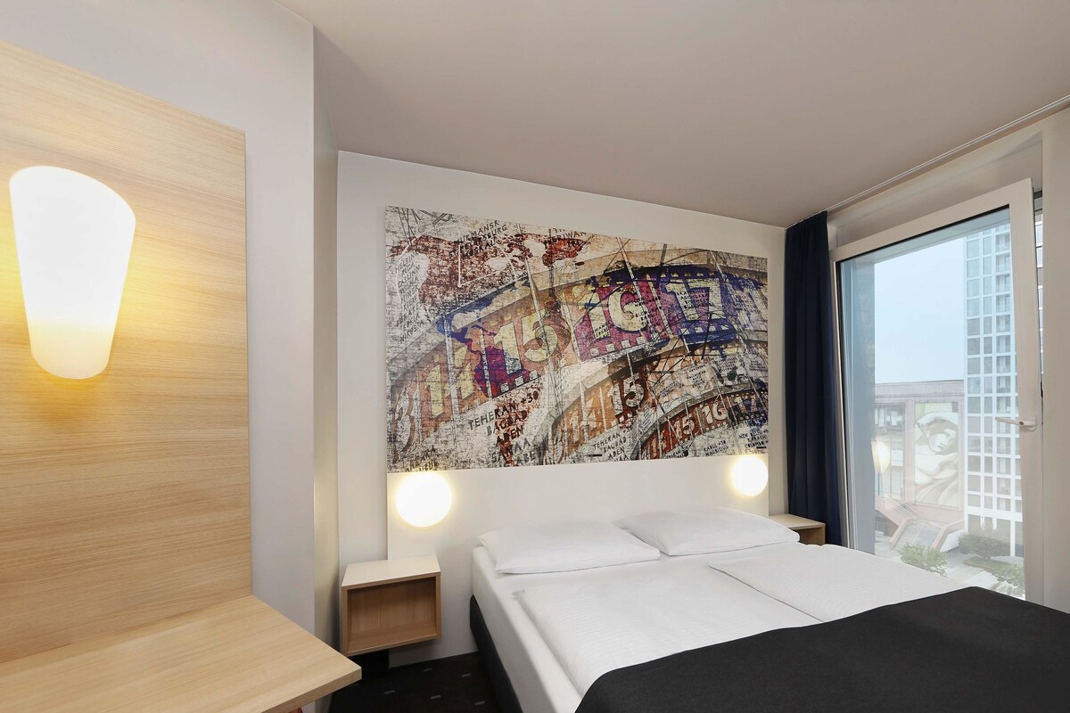 A hotel room with a bed, a wooden nightstand, a wall-mounted lamp, and a large abstract artwork.