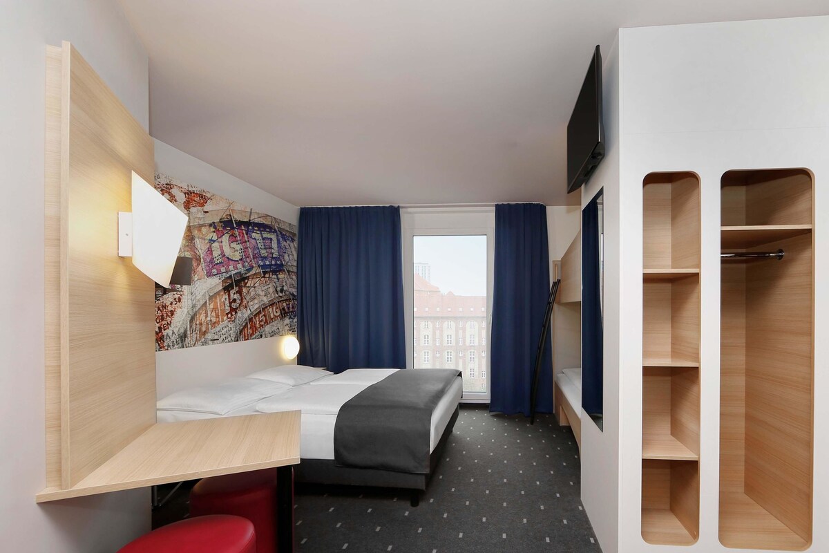 A hotel room with a bed, a desk, a red stool, a wardrobe, a TV, and a cityscape view.