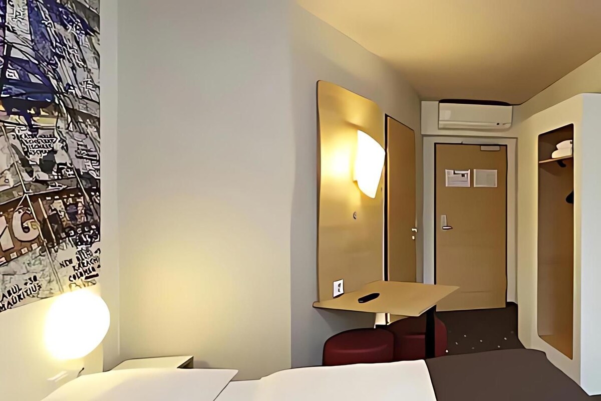 Habitación de hotel con cama, escritorio, silla, espejo y unidad de aire acondicionado.