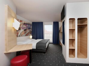 A hotel room with a bed, a desk, a red stool, a wardrobe, a TV, and a cityscape view. - B&B Hotel Berlin-Alexanderplatz (Berlin)