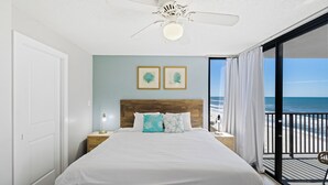 A bedroom with a large bed, a wooden headboard, two framed artworks, a nightstand with a lamp, and a balcony with a view of the beach.