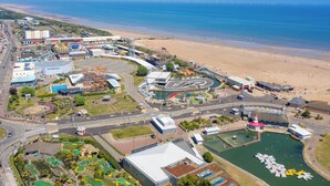 Vue aérienne d’une zone côtière comprenant une plage, un port de plaisance et divers bâtiments, notamment un théâtre et un centre commercial.