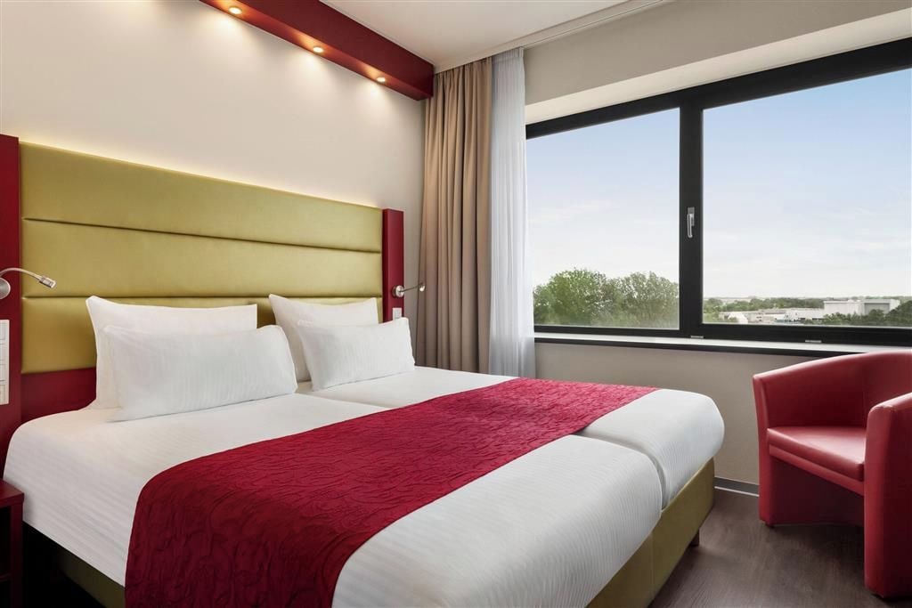 Habitación de hotel con una cama grande, una silla roja y vistas de la ciudad a través de la ventana.