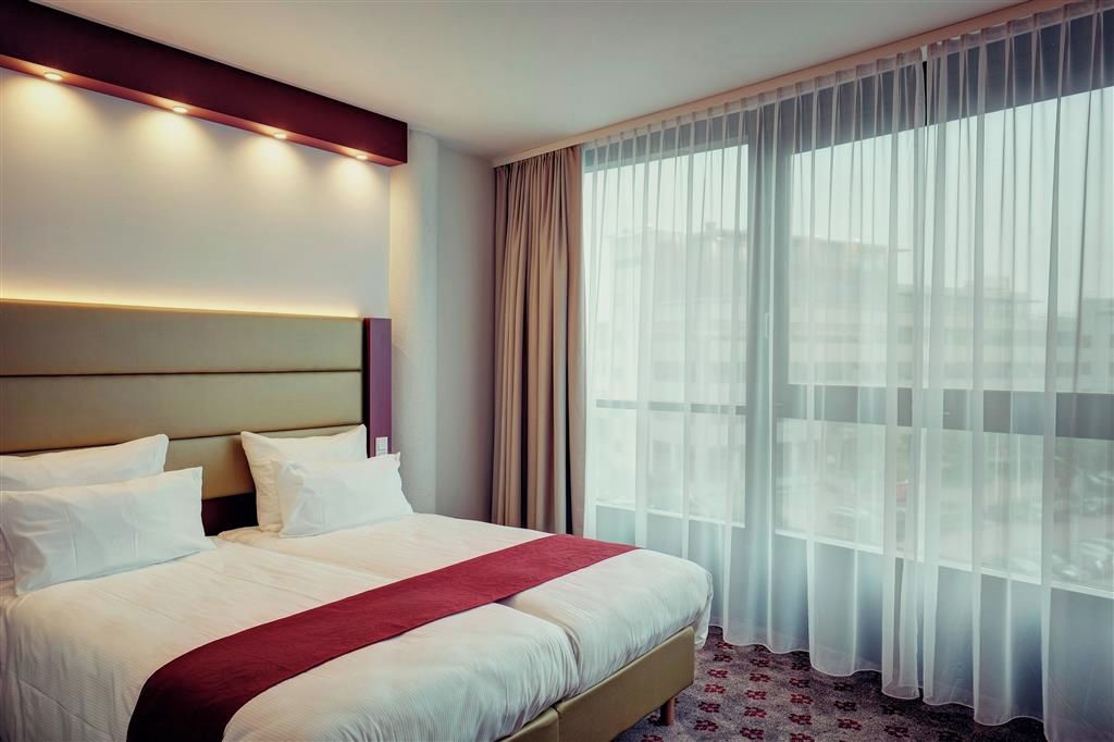 A hotel room with a large bed, a headboard with built-in lighting, and a view through sheer curtains.