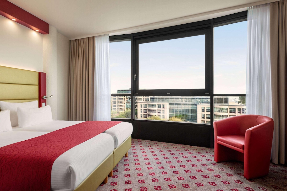 Une chambre d’hôtel avec un lit, un fauteuil rouge, une grande fenêtre et une vue sur les immebles.