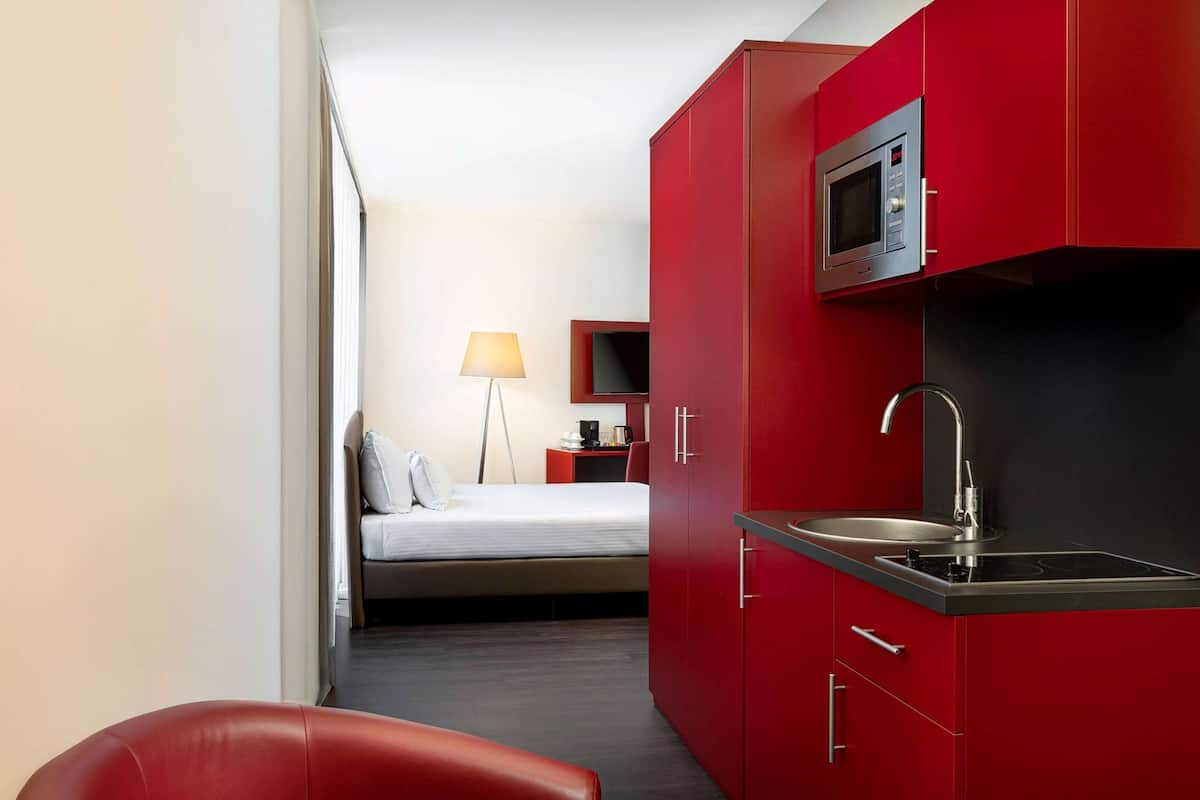 Une chambre d’hôtel compacte comprenant un coin cuisine, un lit, une table de chevet avec une lampe et une chaise rouge.