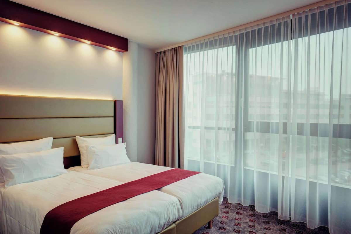 Une chambre d’hôtel dotée d’un grand lit, d’une tête de lit avec éclairage intégré, d’une fenêtre avec des rideaux légers et d’un sol recouvert de moquette.