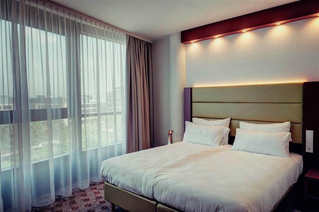 Een hotelkamer met een groot bed, een nachtkastje en een raam met dunne gordijnen.