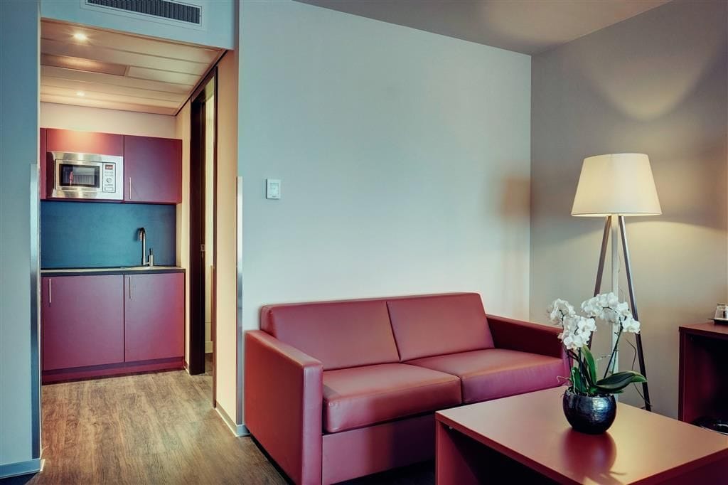 Una sala moderna con un sofá rojo, una mesita y una lámpara. Hay una zona de cocina con microondas y armarios.
