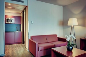 Een moderne woonkamer met een rode bank, een kleine tafel en een lamp. Er is een keuken met een magnetron en kastjes.