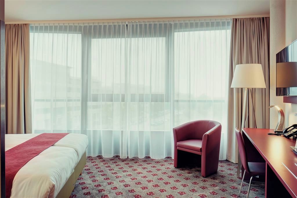 Quarto de hotel com cama, escrivaninha, cadeira e uma grande janela com cortinas leves.