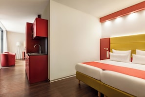 A modern hotel room with a bed, a red minibar, and a small kitchen area.